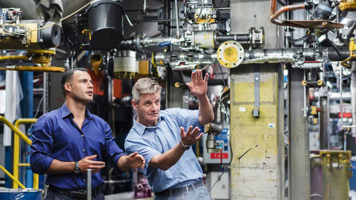 SIFA Fachkraft für Arbeitssicherheit Zwei Fachkräfte für Arbeitssicherheit beim Gespräch in einer großen Industriehalle