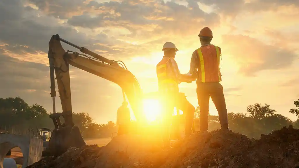 Zwei Männer auf einer Baustelle Ihres Unternehmens blicken in der Abenddämmerung auf Ihre Baupläne