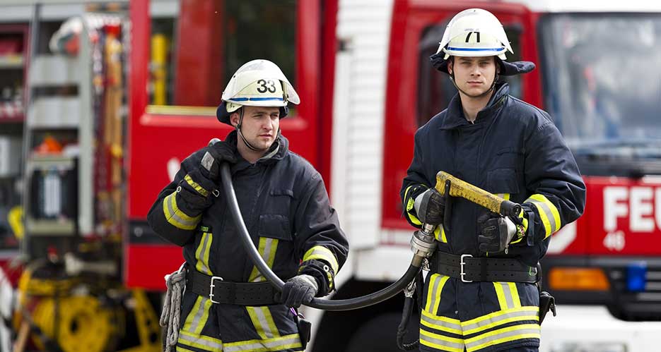 Zwei Feuerwehrleute tragen einen Schlauch zum Einsatzort währedn im Hintergrund der Einsatzwagen steht